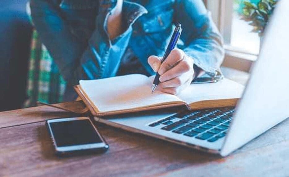 Person writing in a notebook beside a laptop and smartphone